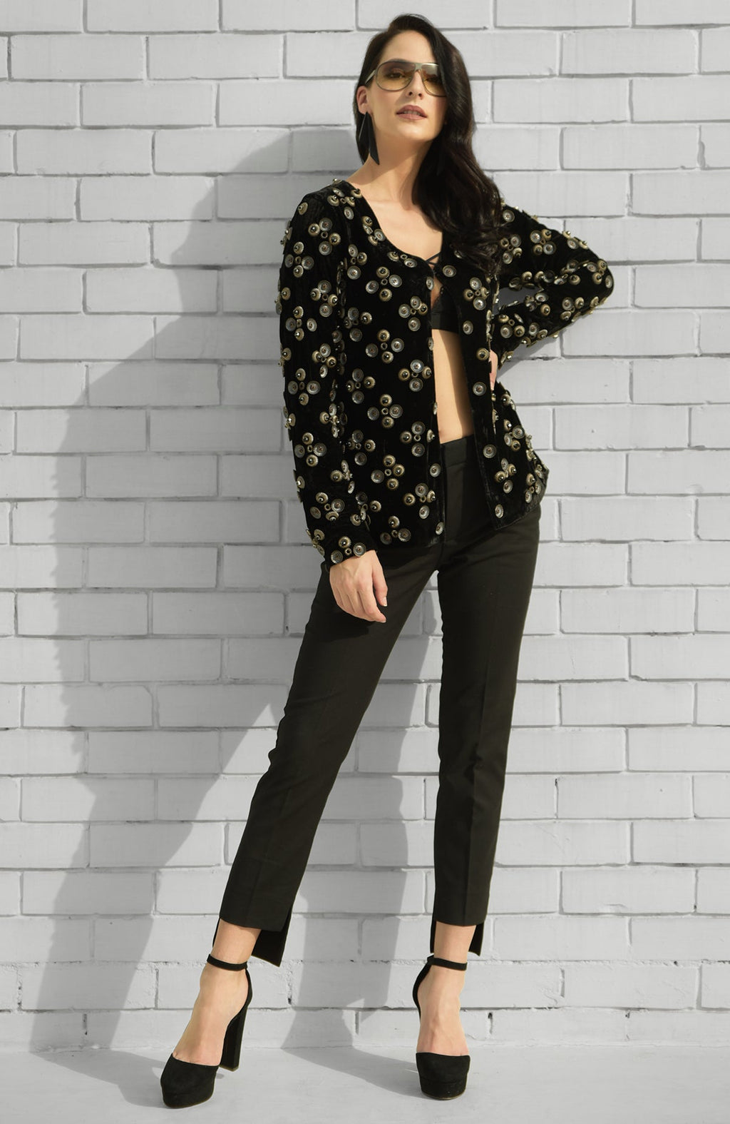 A woman models a black velvet jacket with metal studs, black trousers, and heels.