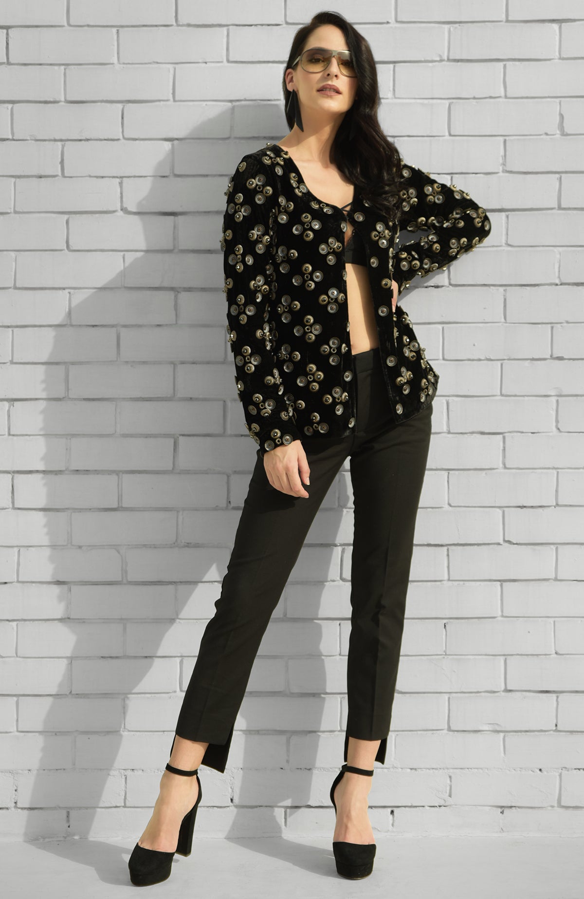 A woman models a black velvet jacket with metal studs, black trousers, and heels.
