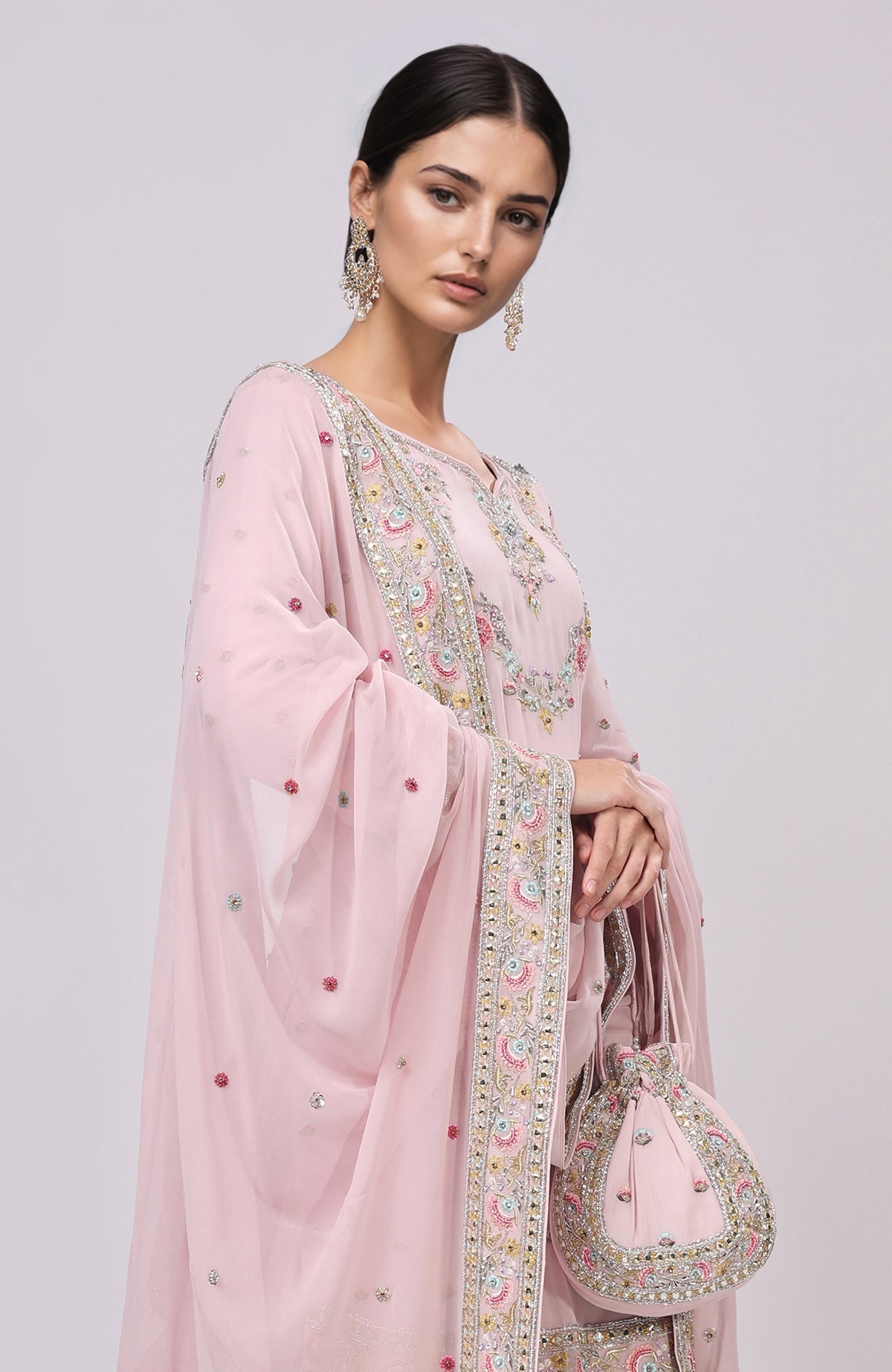 A woman models a marshmallow pink sharara set with intricate embroidery and a matching potli bag.