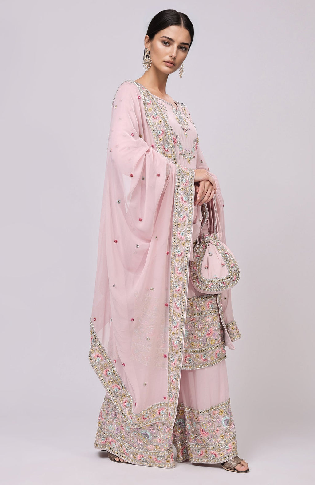A woman models a marshmallow pink sharara set with intricate embroidery and a matching potli bag.