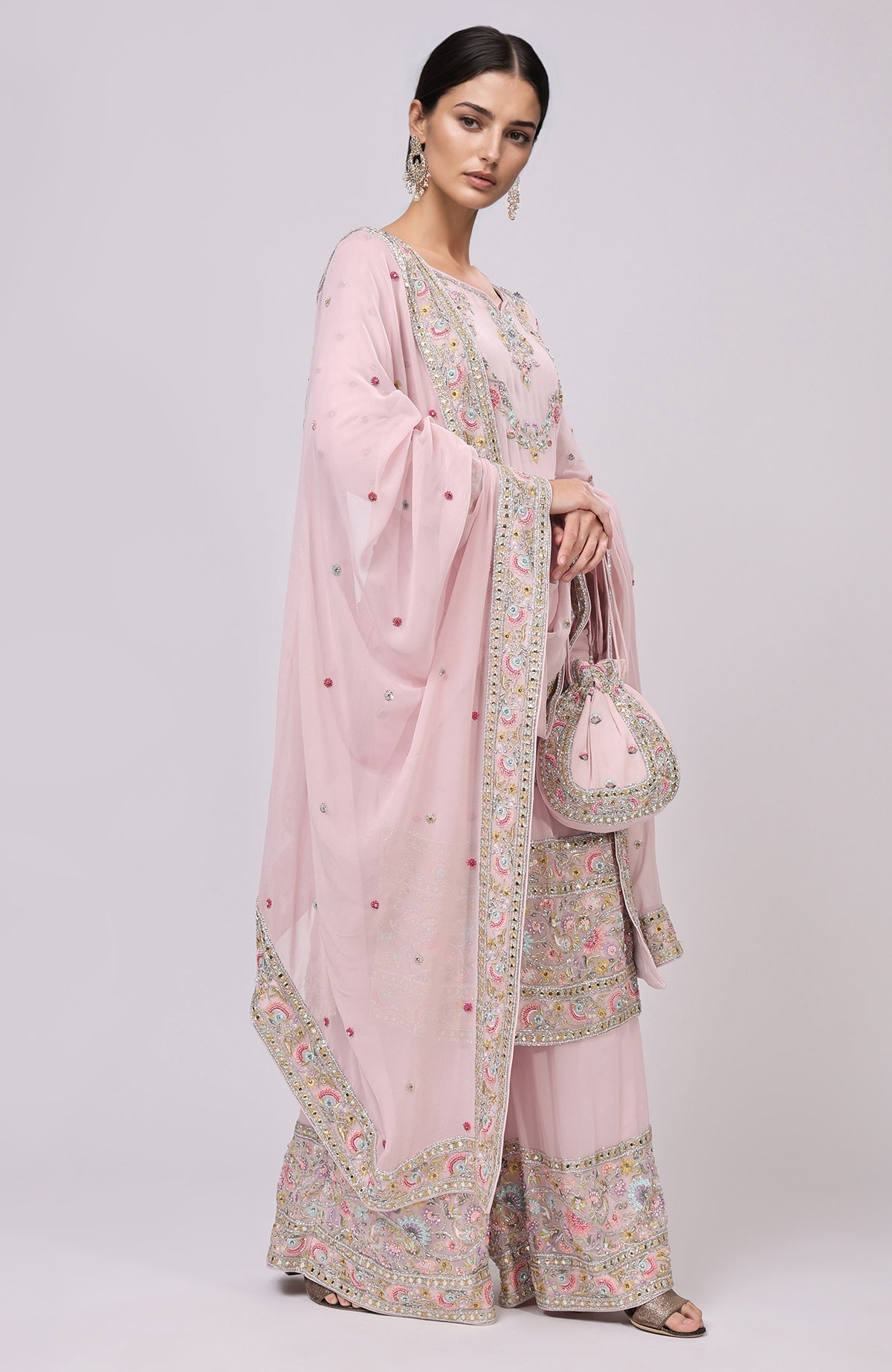 A woman models a marshmallow pink sharara set with intricate embroidery and a matching potli bag.