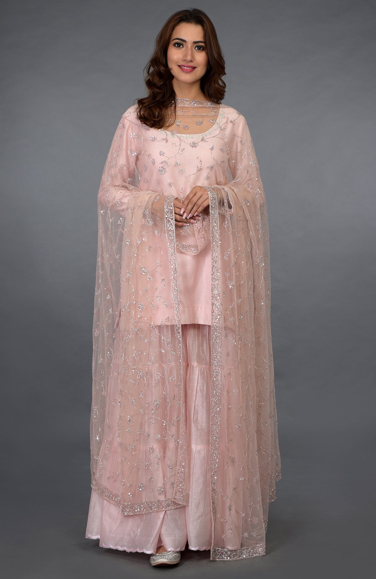 A woman models a pink gharara suit with silver zari, beads, and sequin embroidery.