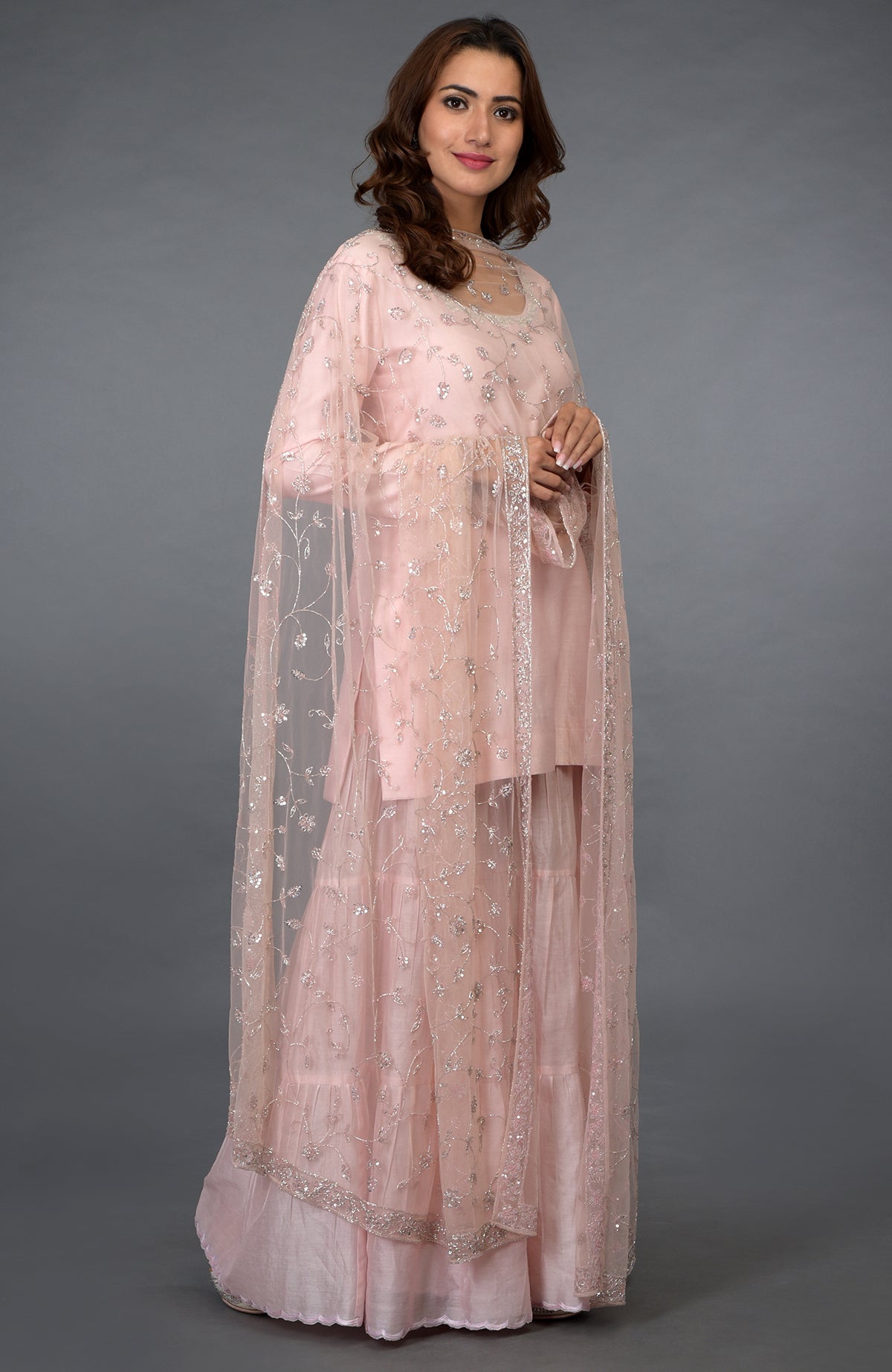 A woman models a pink gharara suit with silver zari, beads, and sequin embroidery.