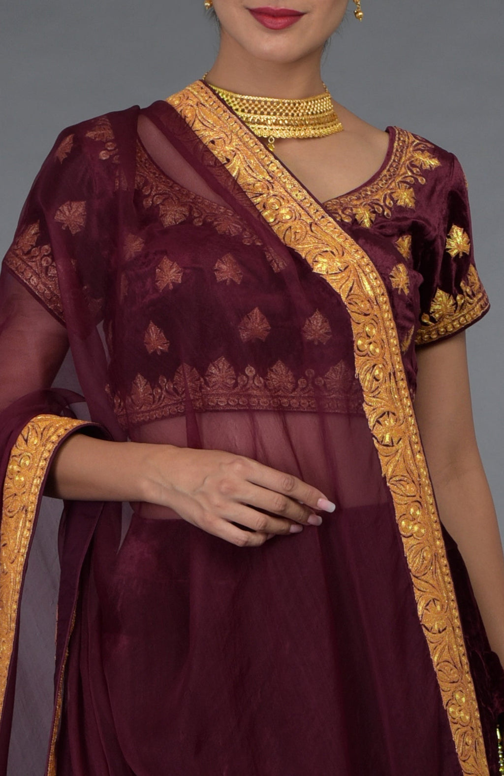 A woman models a burgundy Kashmiri Tilla embroidered lehenga outfit with gold jewelry.