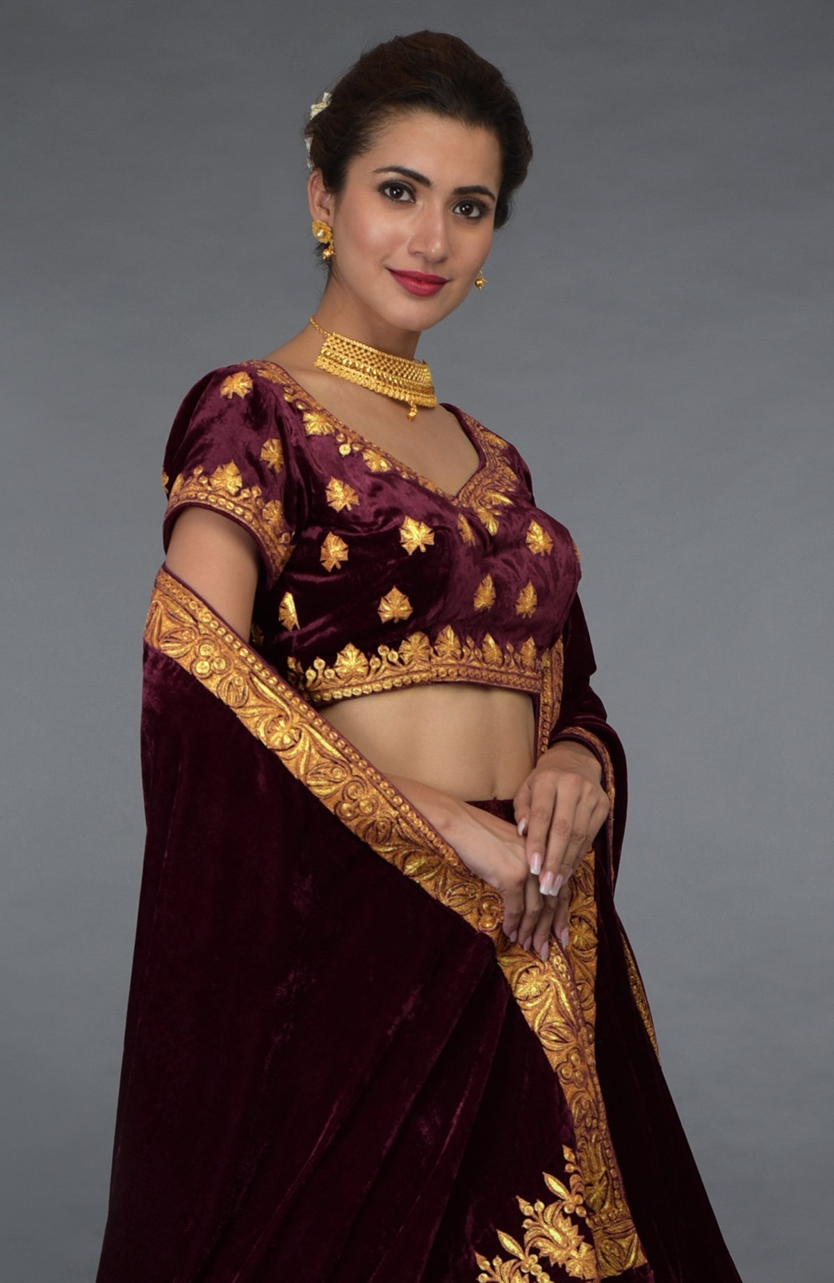A woman models a burgundy velvet lehenga outfit with gold tilla embroidery.