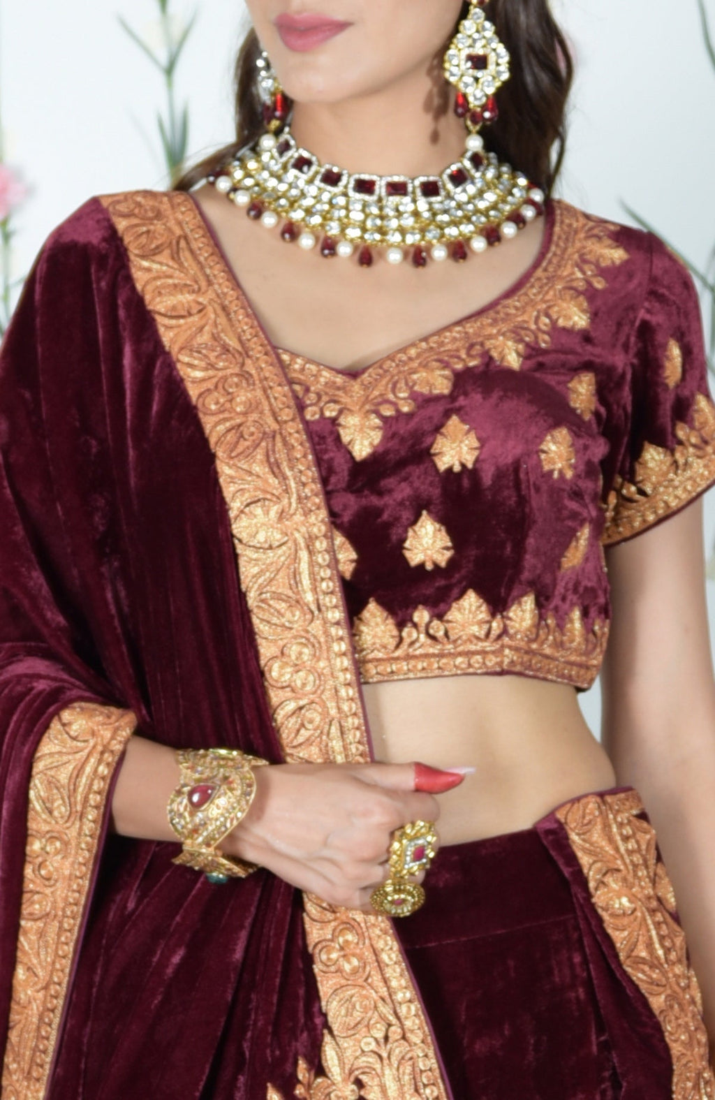 A woman models a burgundy velvet lehenga with gold tilla embroidery and matching jewelry.