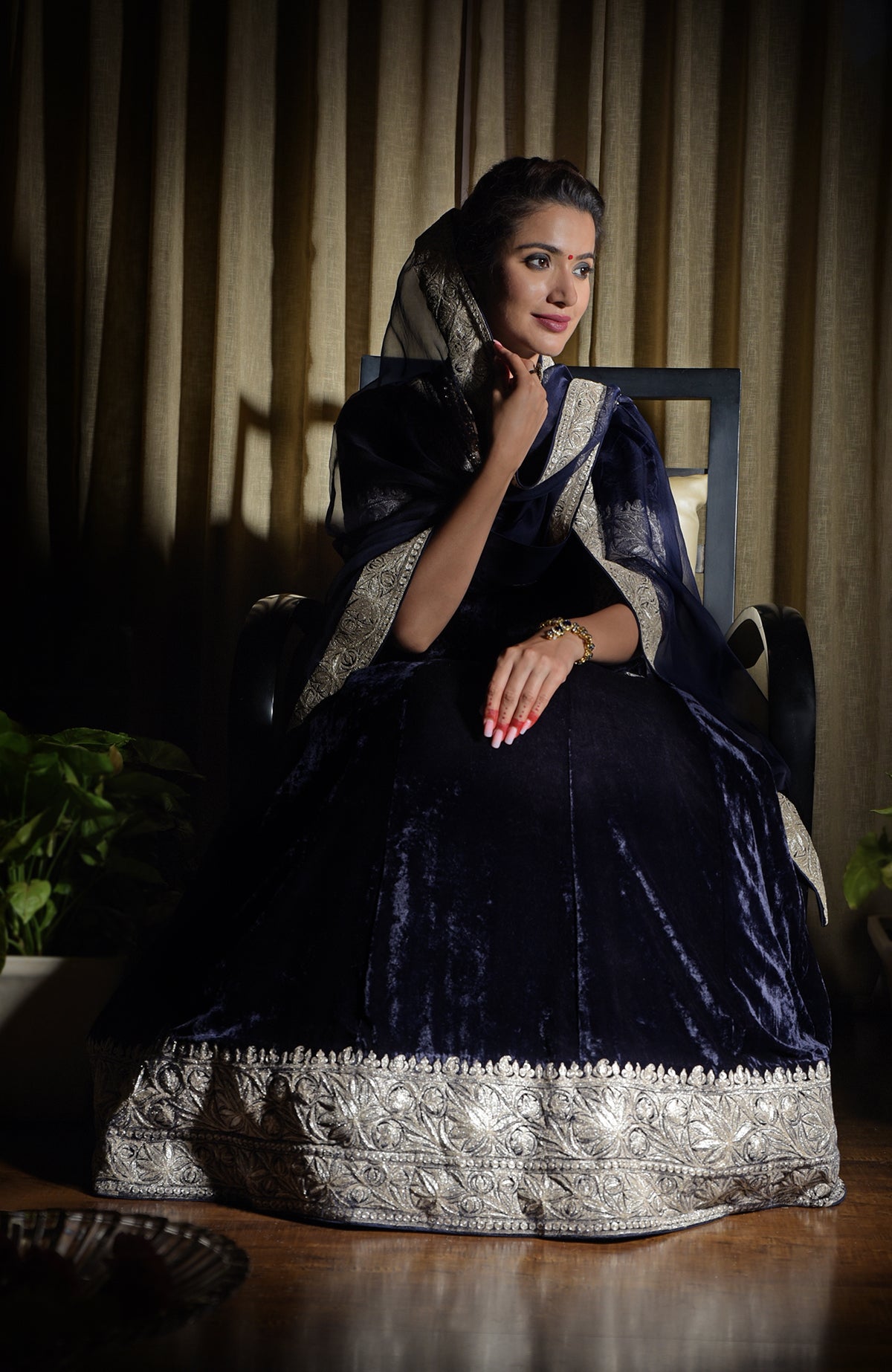 A woman in a midnight blue velvet lehenga with silver tilla embroidery sits on a chair.