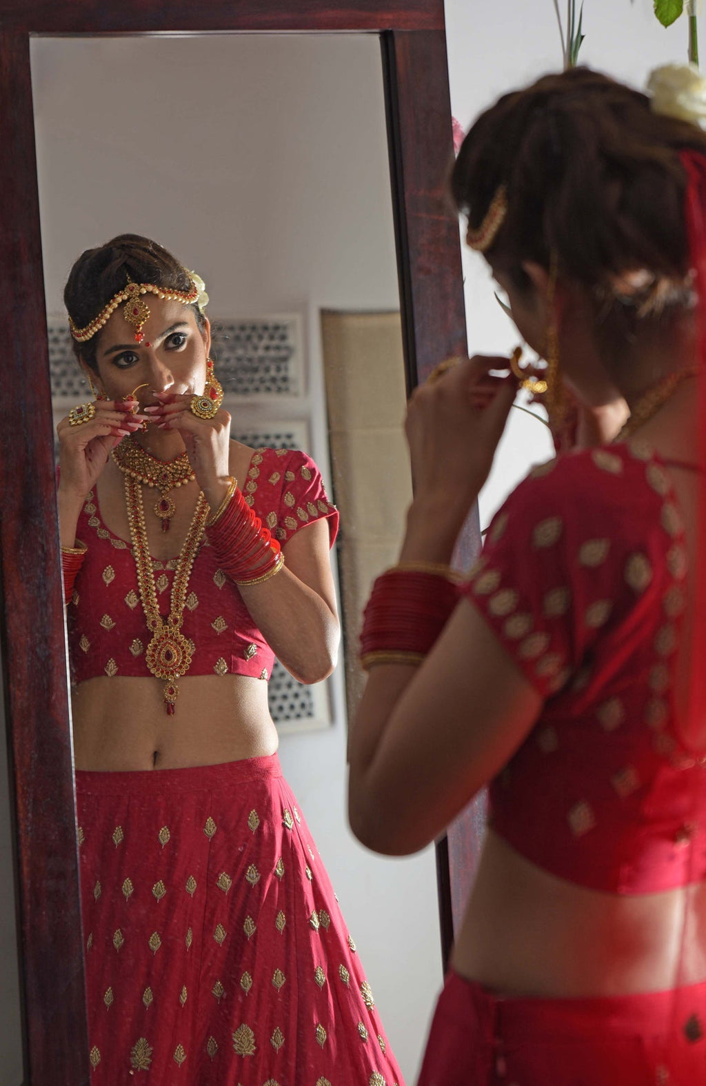 A woman in a red embroidered lehenga and jewelry adjusts her nose ring in a mirror.