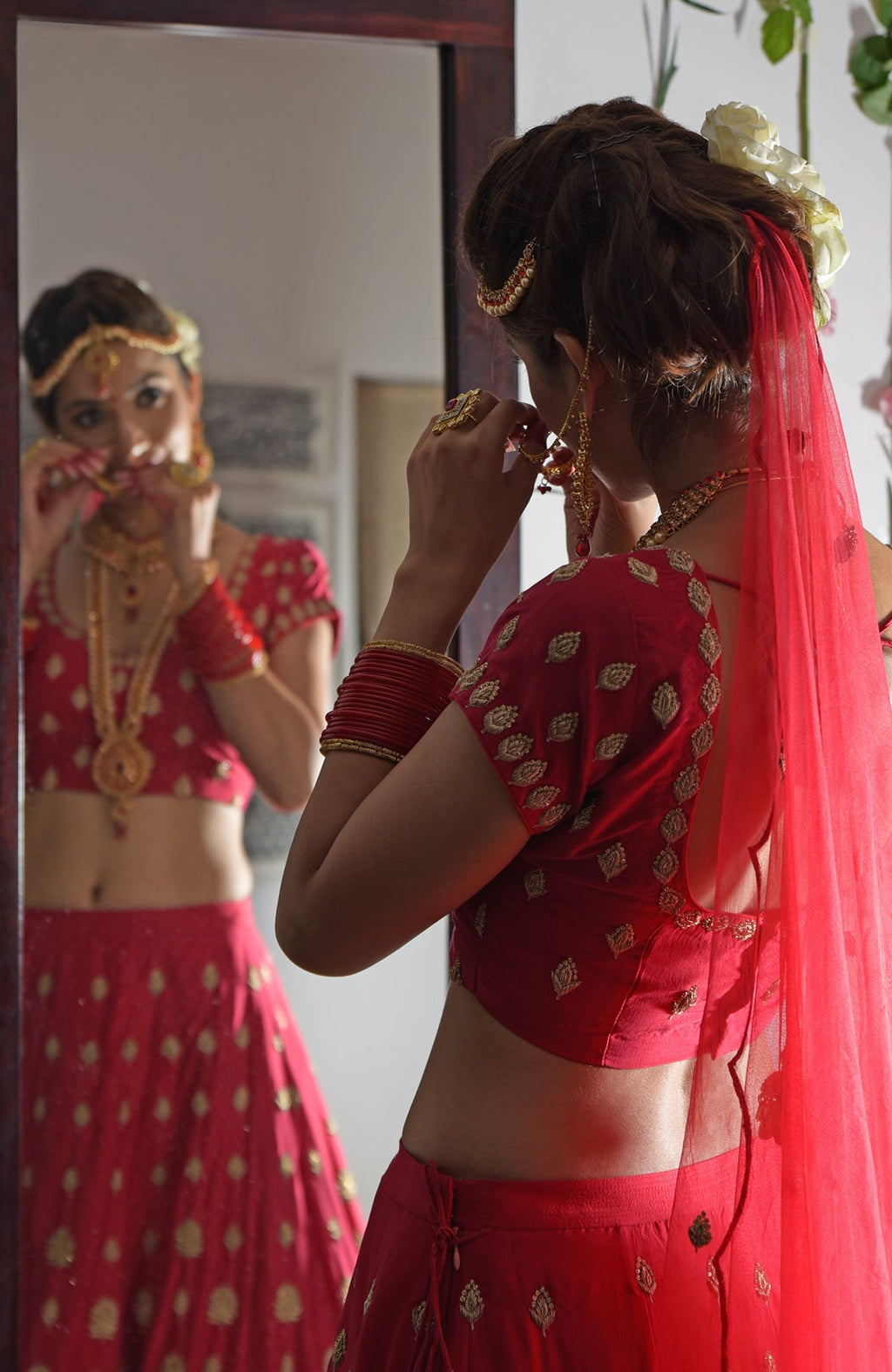 A woman in a red Zardozi and crystal embroidered lehenga outfit looks in a mirror.