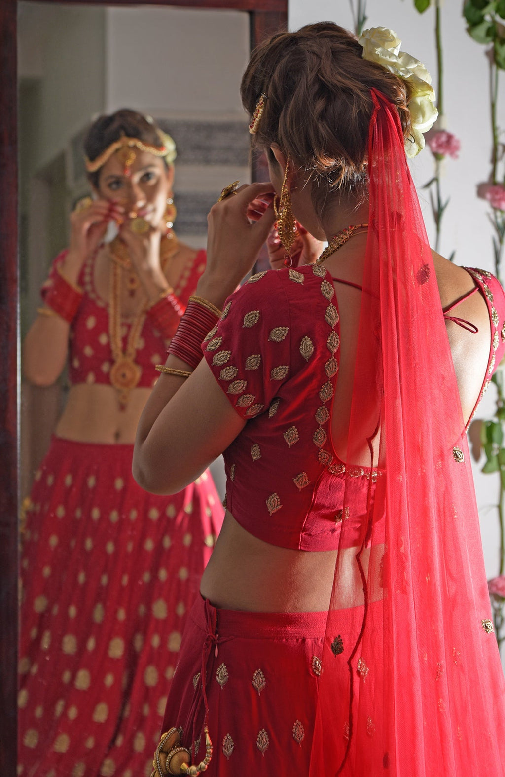 A woman in a red Zardozi and crystal embroidered lehenga adjusts her earring in a mirror.