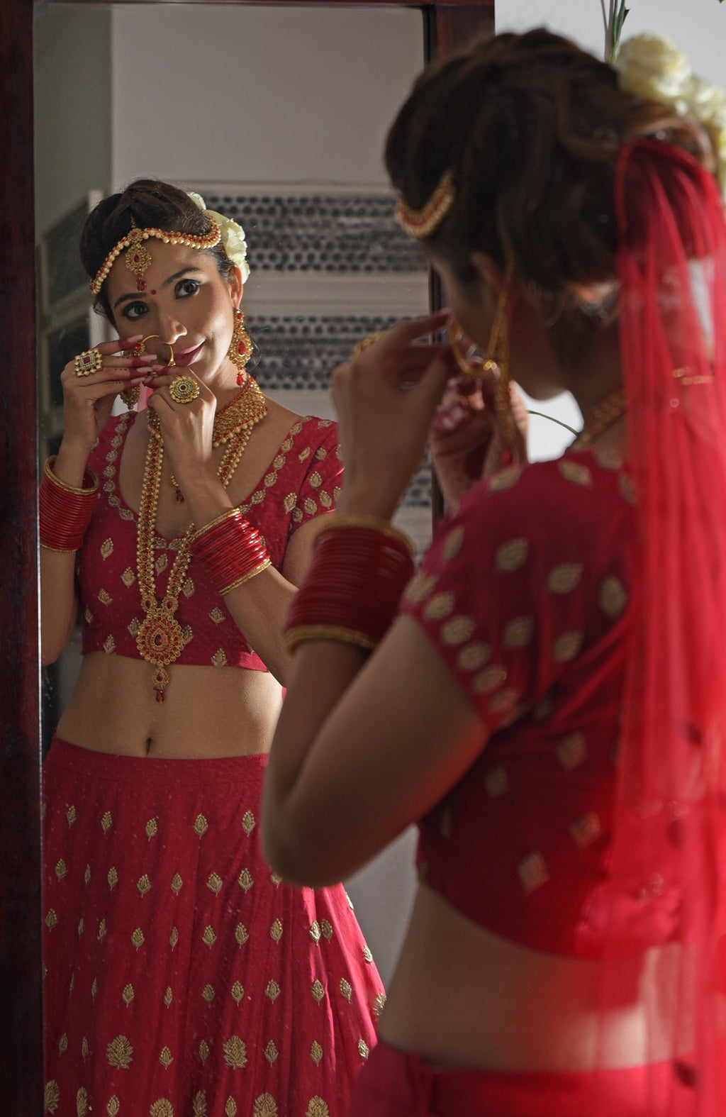 A woman in a red embroidered lehenga and jewelry adjusts her nose ring in a mirror.