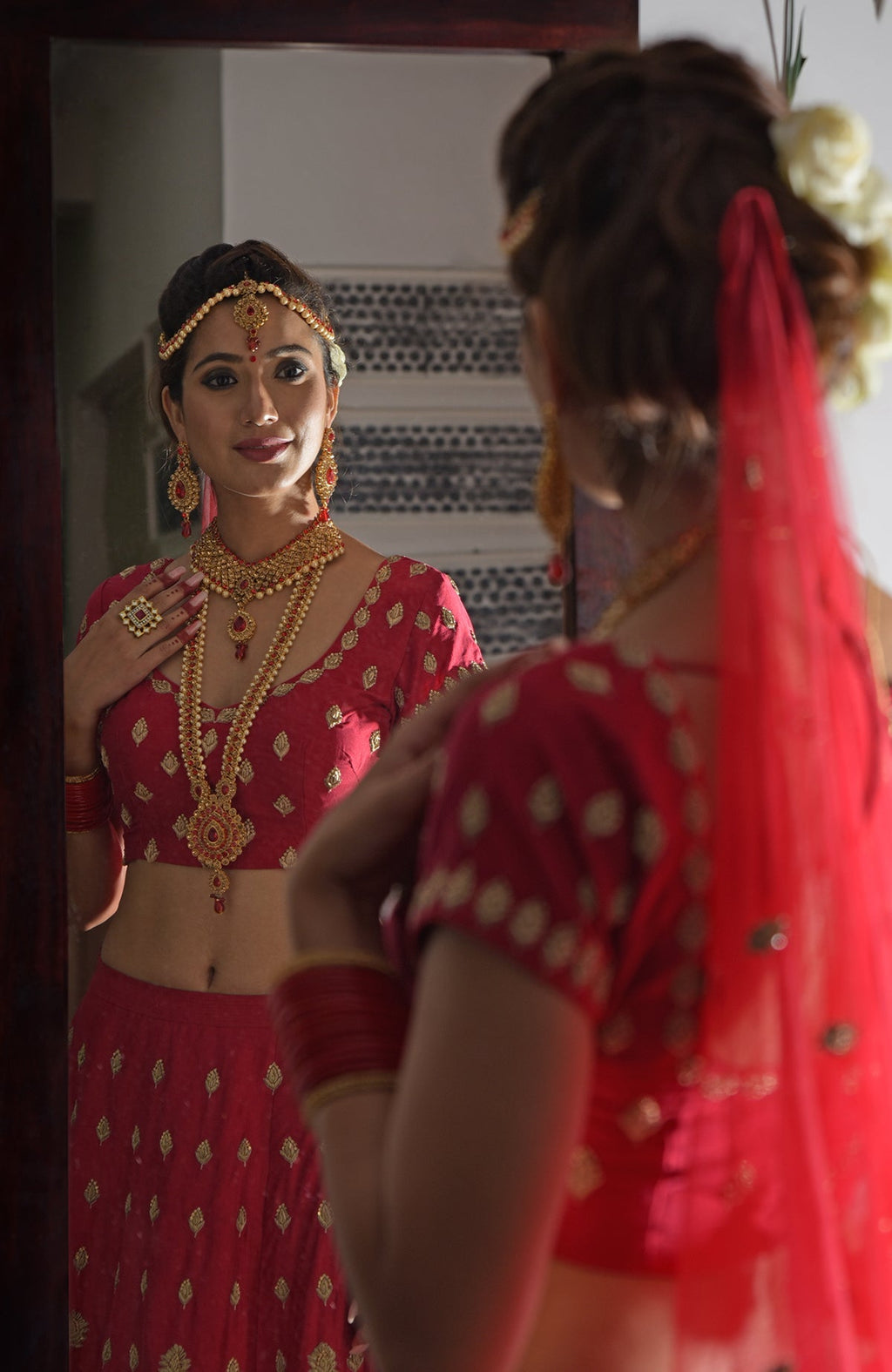 A woman in a red and gold embroidered lehenga outfit looks at her reflection in a mirror.