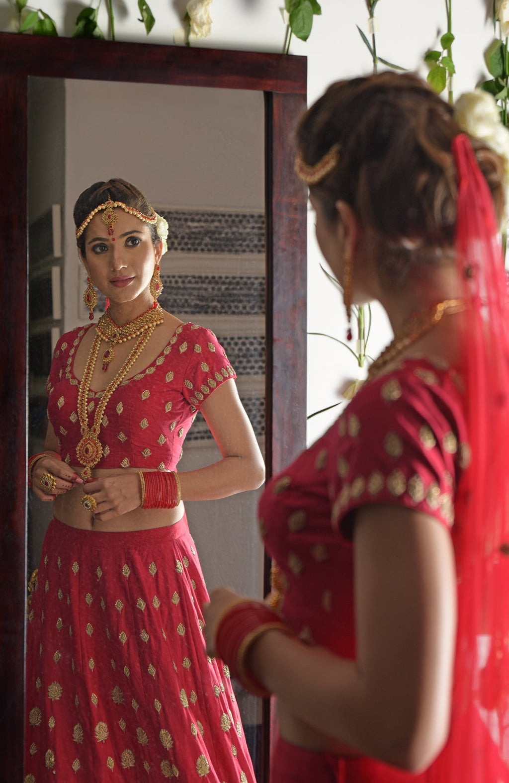 A woman in a red and gold embroidered lehenga outfit looks in a mirror.
