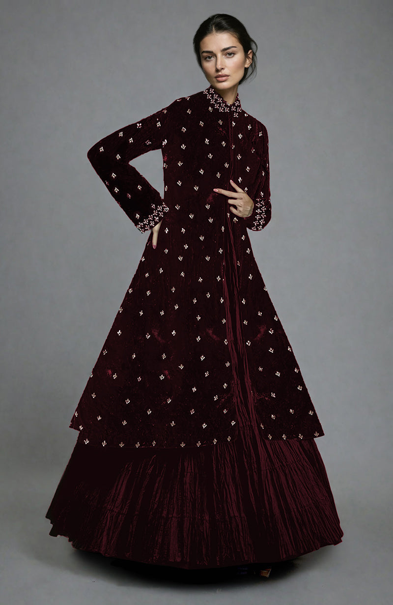 A model wears a long, maroon velvet jacket with gold embroidery over a matching skirt.