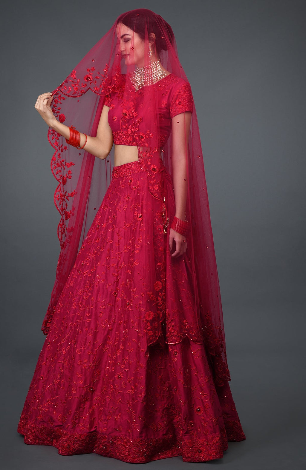 A woman models a red floral embroidered lehenga with a matching veil and jewelry.