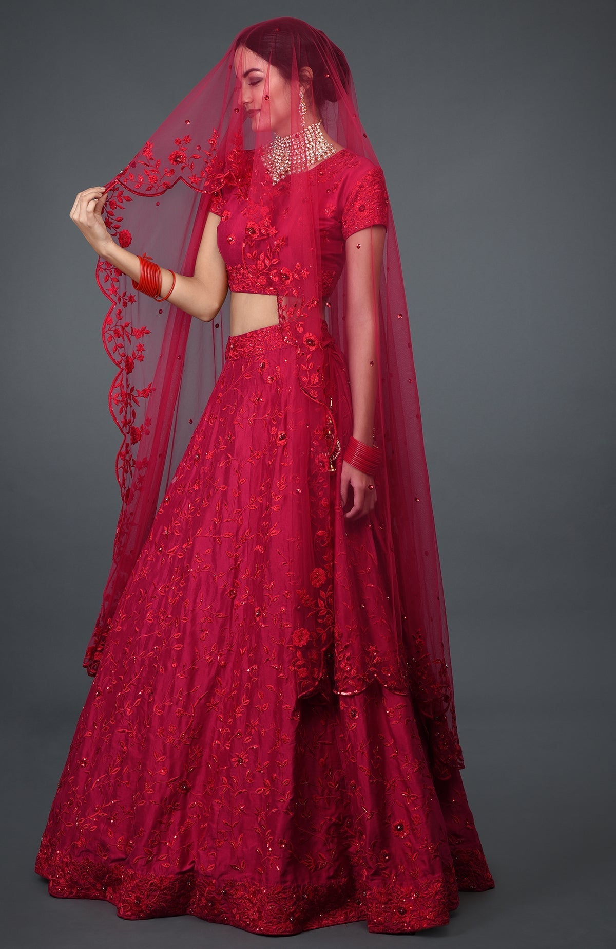 A woman models a red floral embroidered lehenga with a matching veil and jewelry.