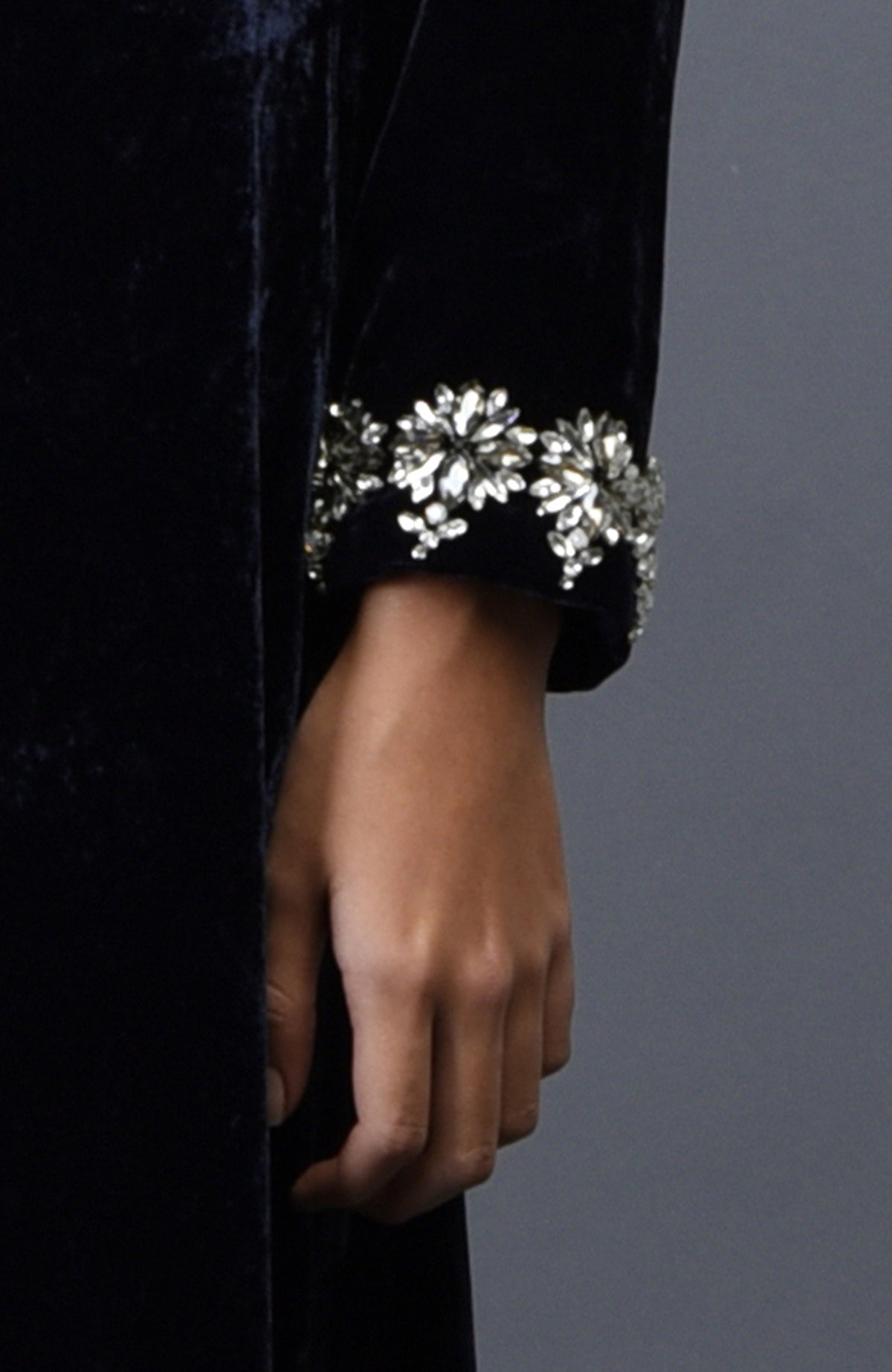 A woman's hand rests against a midnight blue velvet sleeve with crystal floral embroidery.