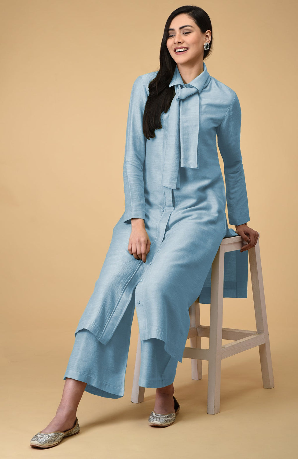 A woman in a light blue tie-neck kurta and wide-leg pants sits on a stool.