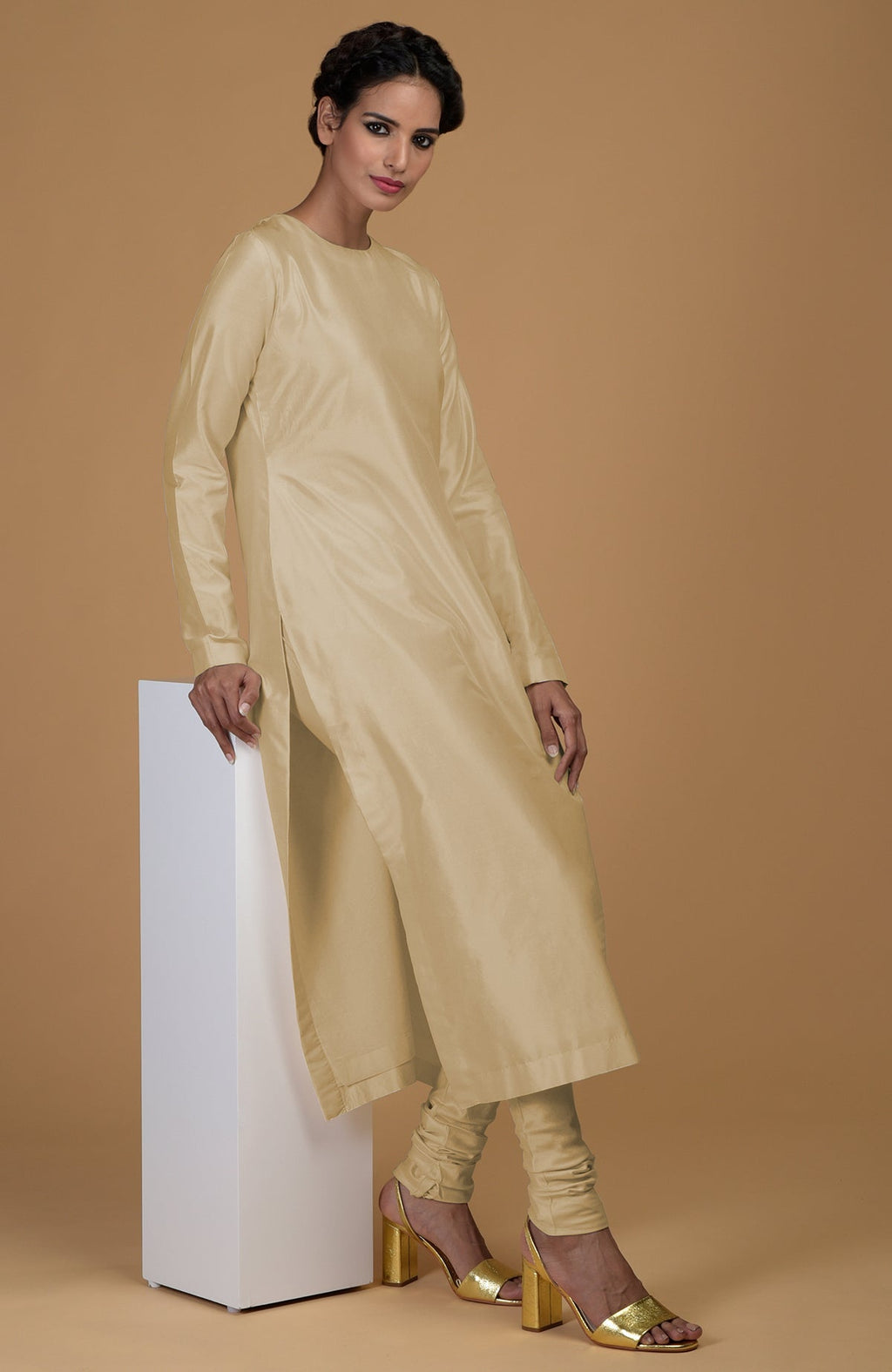 A woman in a beige pure Matka silk kurta set leans on a white pedestal.