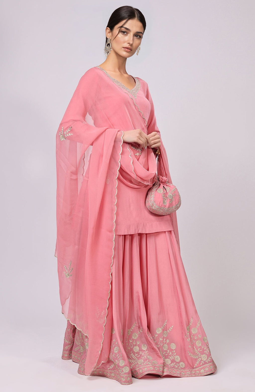 A woman models a pink sharara set with gold embroidery and a matching potli bag.
