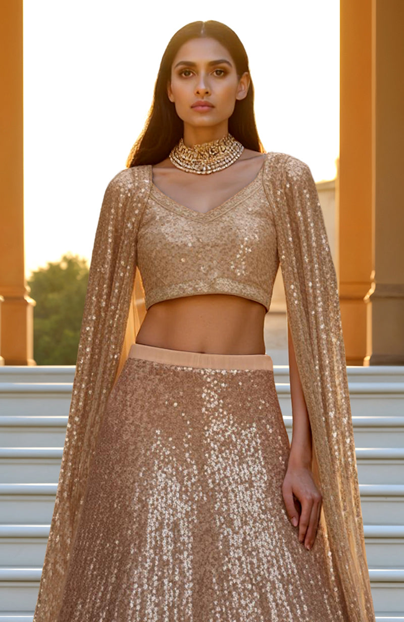 Champagne gold lehenga set with sequin embroidery, cropped blouse, and flowing cape sleeves.