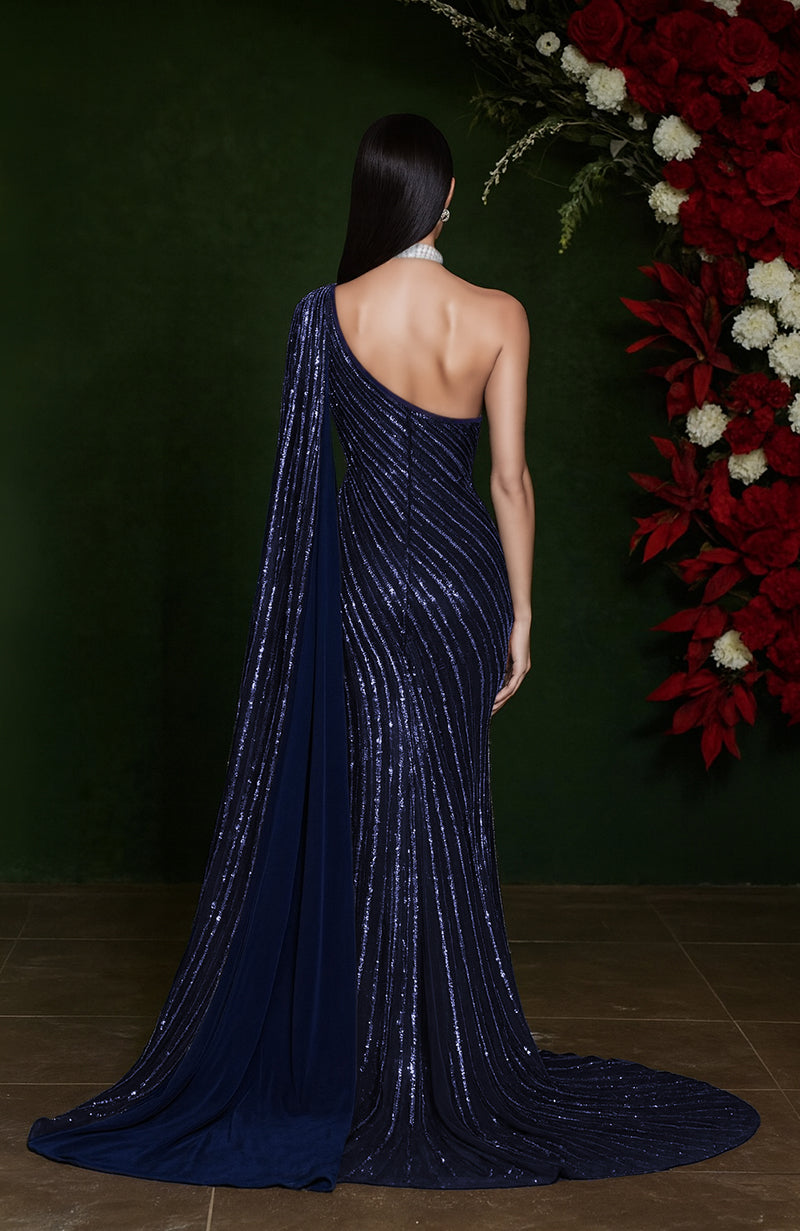 One-shoulder midnight blue gown with crystal beadwork, draped cape, and thigh-high slit.