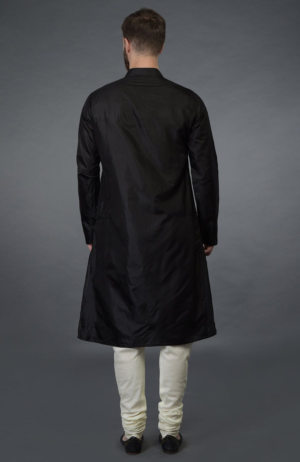 A man from behind wearing a black silk kurta and cream churidar pants.