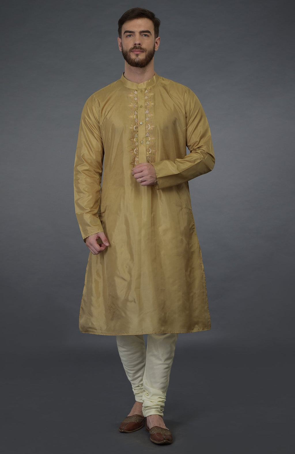 A man models a gold embroidered silk kurta set with ivory churidars and brown shoes.