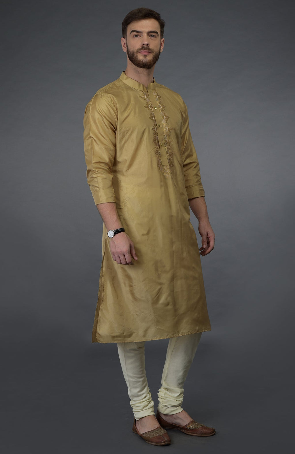 A man models a gold embroidered silk kurta set with ivory churidars and brown juttis.