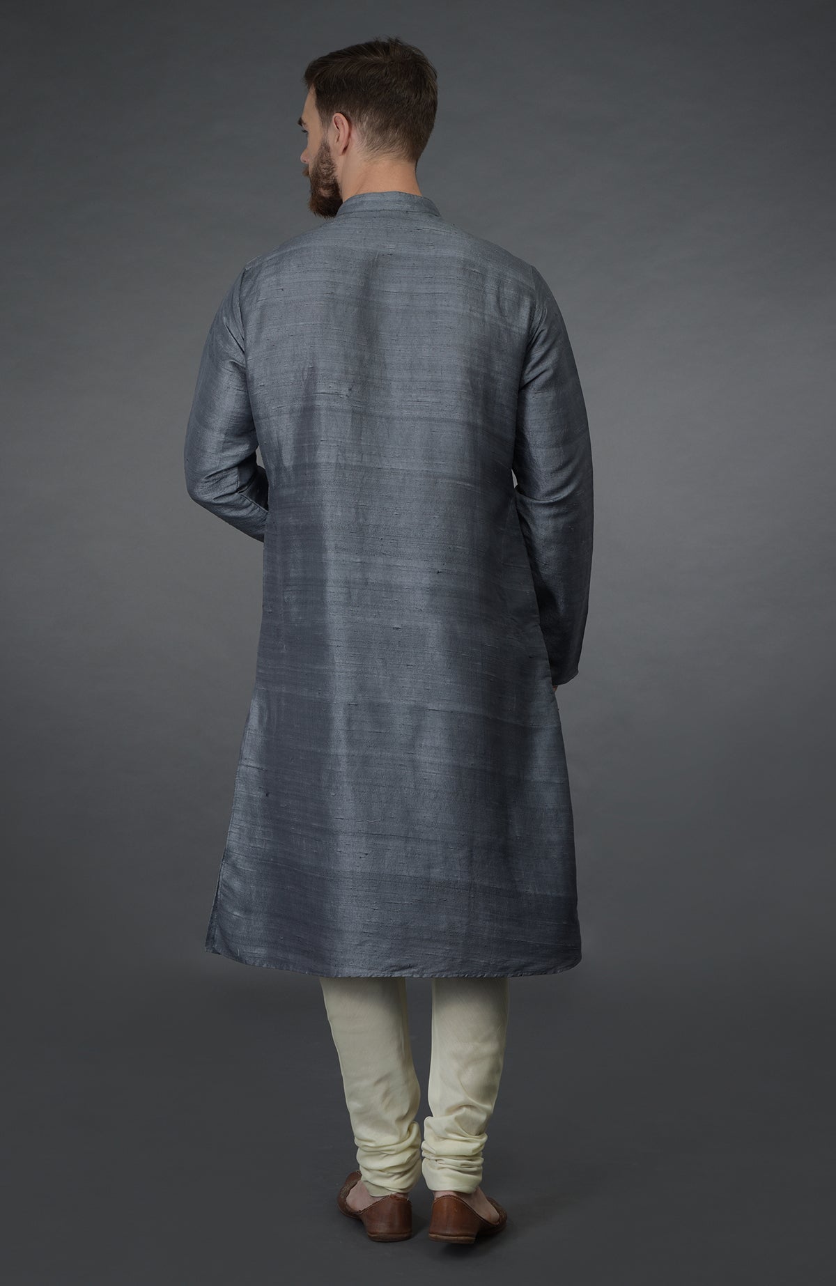 A man from behind wears a grey silk kurta and ivory churidar pants with brown shoes.