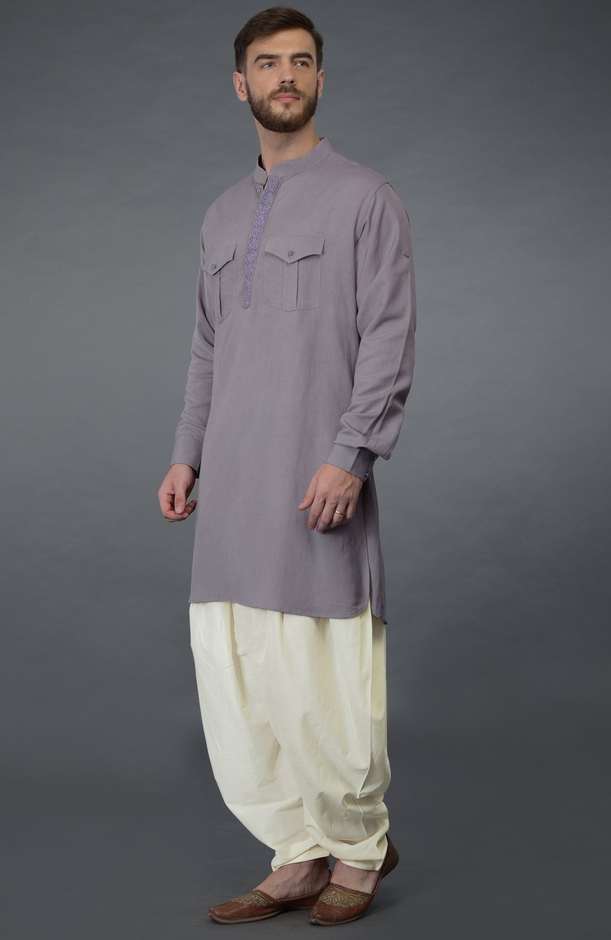 A man wears a lilac grey embroidered linen kurta with ivory pathani salwar pants and brown shoes.