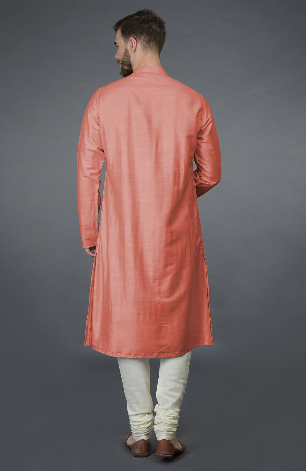 A man from behind wears a terracotta kurta and ivory churidar pants with brown shoes.