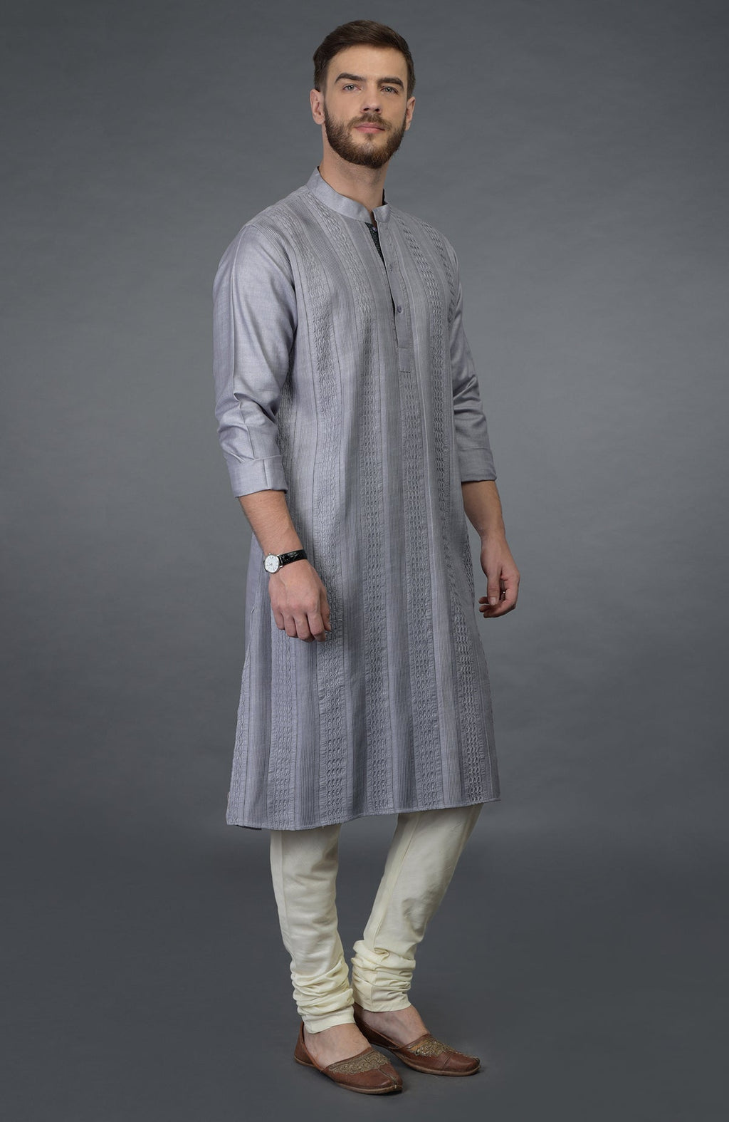 A man models a grey pintuck silk kurta set with ivory churidar pants and brown shoes.