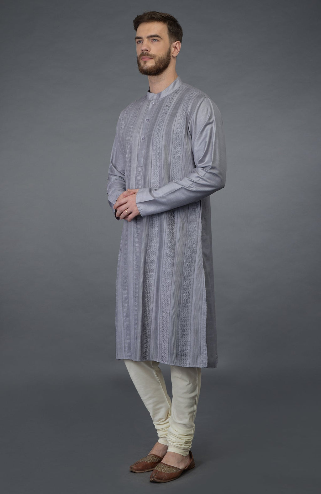 A man models a grey pintuck silk kurta set with ivory churidar pants and brown shoes.