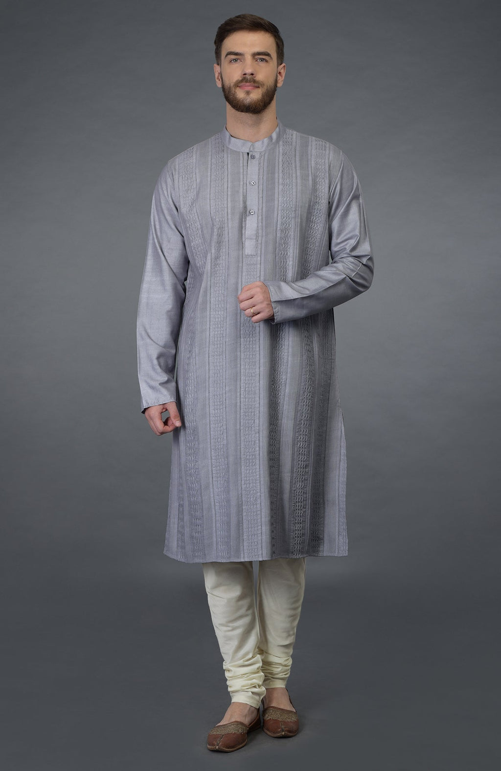 A man models a grey pintuck silk kurta set with ivory churidar pants and brown shoes.