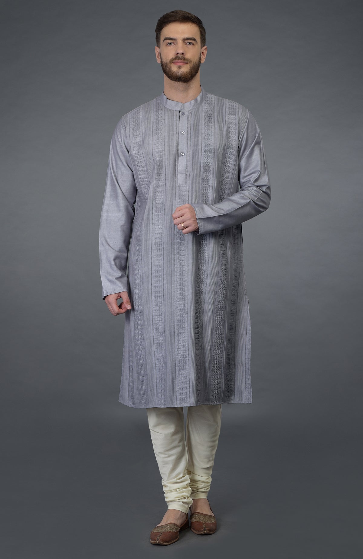 A man models a grey pintuck silk kurta set with ivory churidar pants and brown shoes.