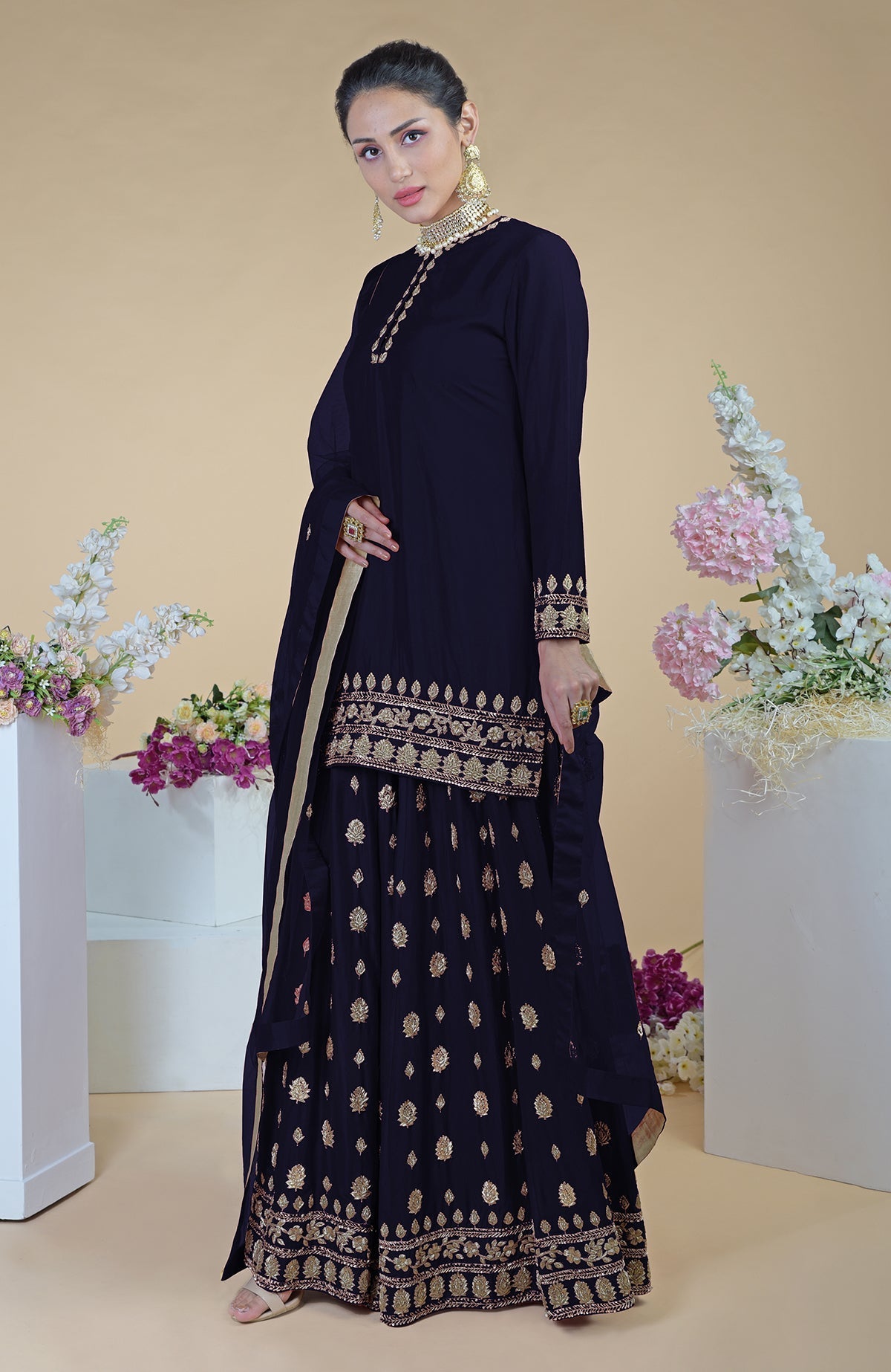 A woman models a navy blue sharara set with gold embroidery and a pearl choker.
