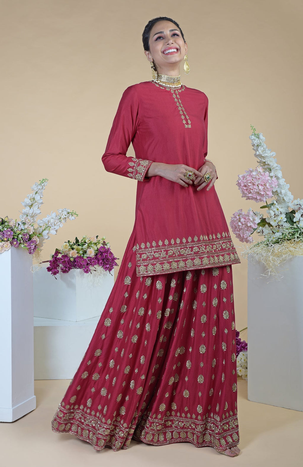 A woman in a crimson-gold zardozi dabka hand-embroidered sharara set smiles.