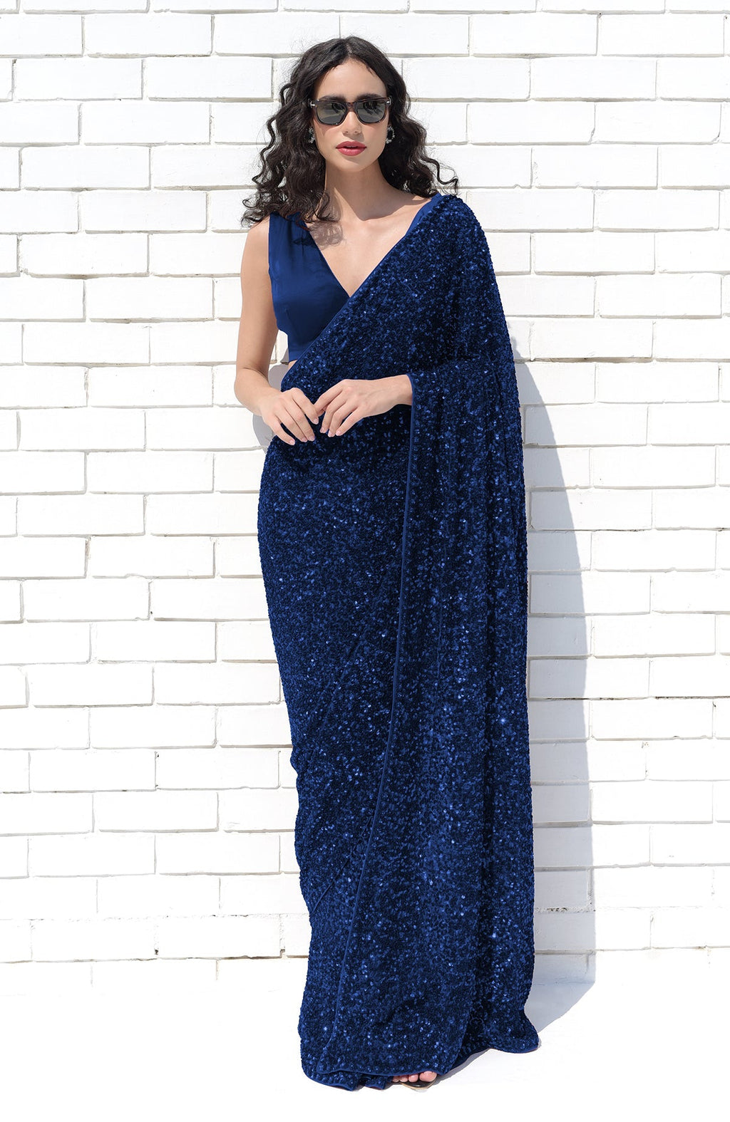 A woman in a sapphire blue sequin saree and matching blouse stands against a white brick wall.