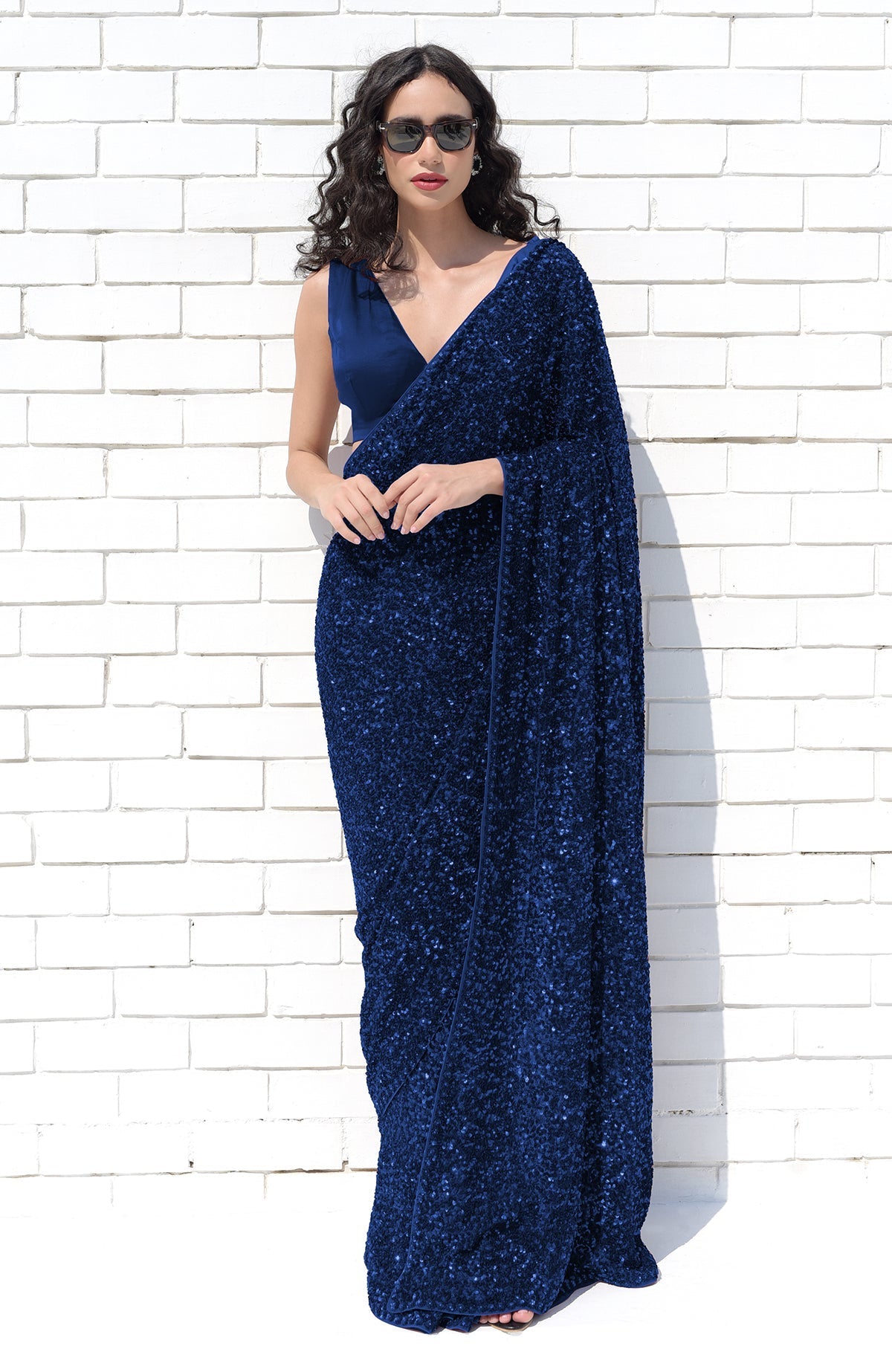 A woman in a sapphire blue sequin saree and matching blouse stands against a white brick wall.