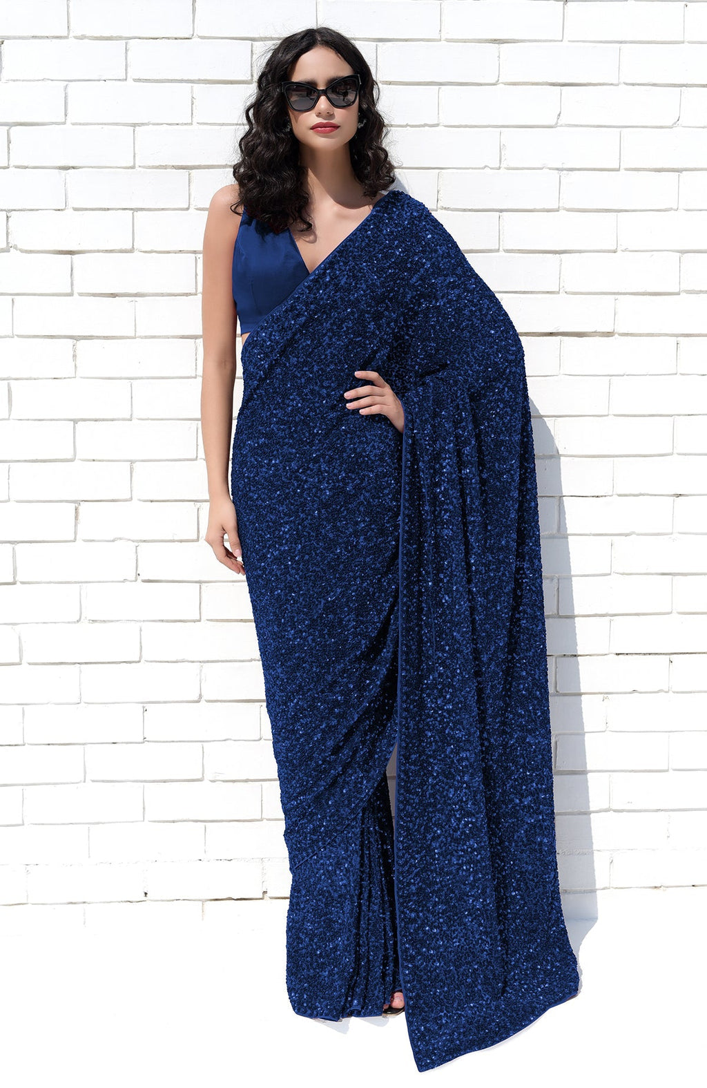 A woman in a sapphire blue sequin saree and matching blouse stands against a white brick wall.
