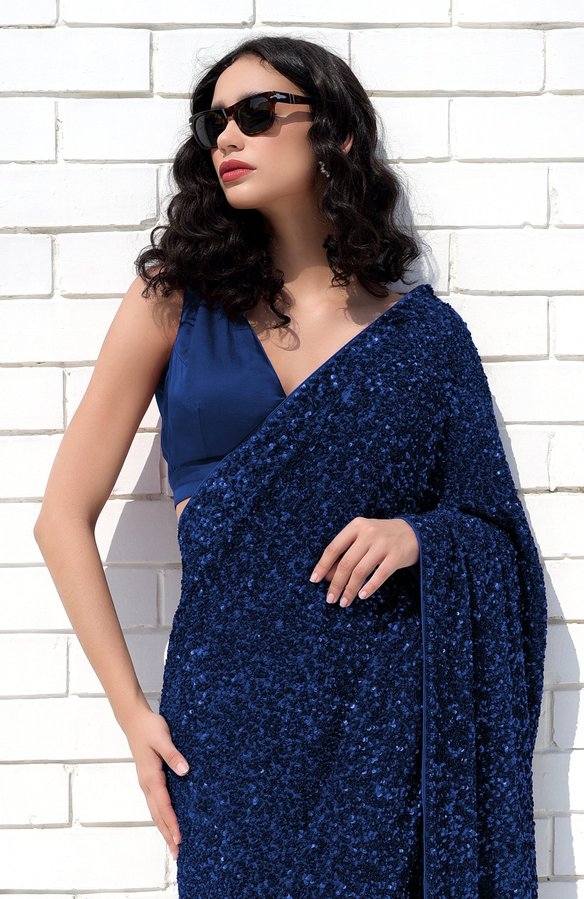 A woman in a sapphire blue sequin saree and matching blouse poses against a white brick wall.