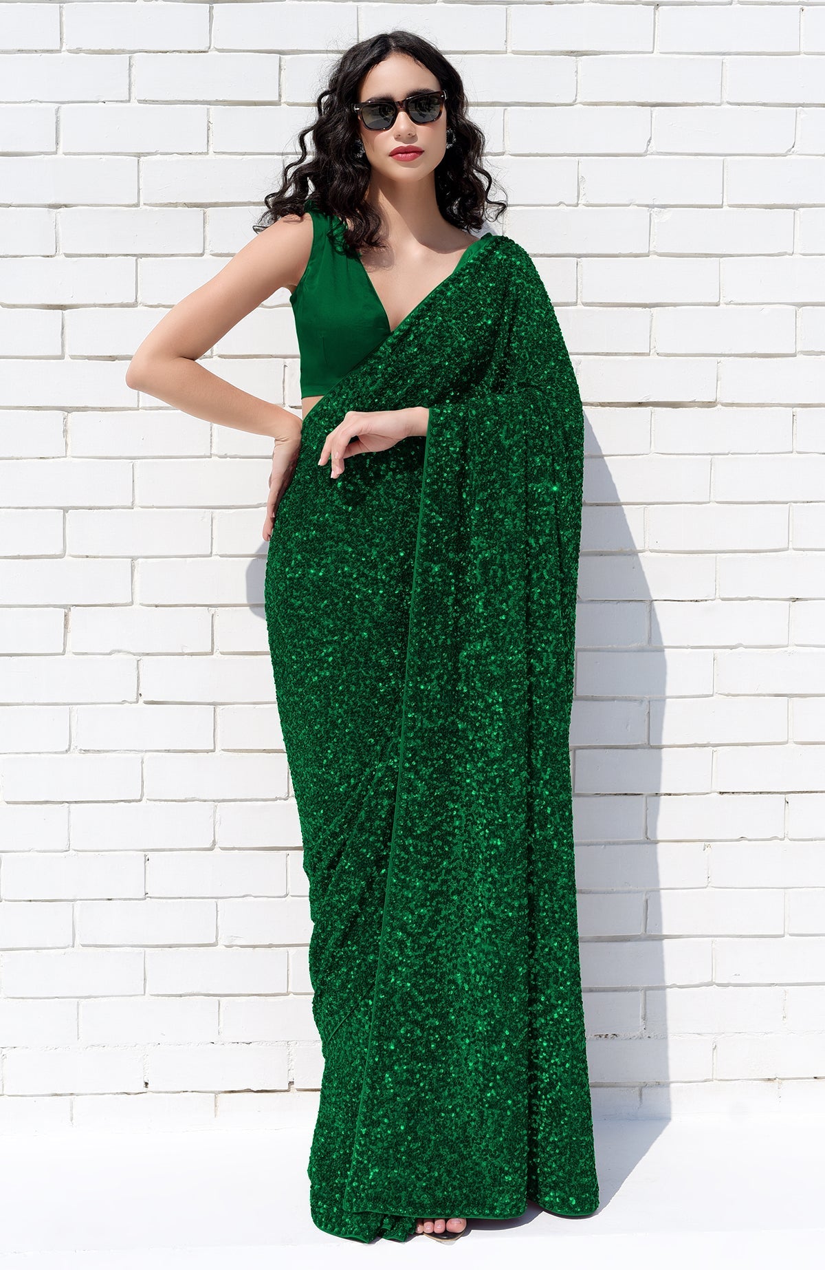 A woman in an emerald green sequin saree and matching blouse poses against a white brick wall.