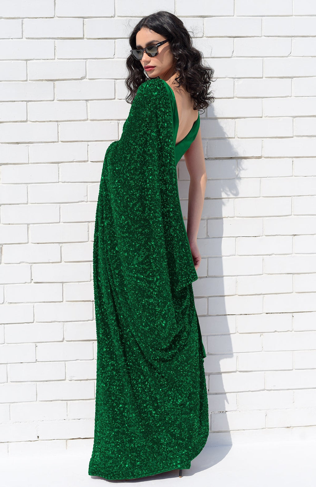 A woman in an emerald green sequin saree and sunglasses poses against a white brick wall.