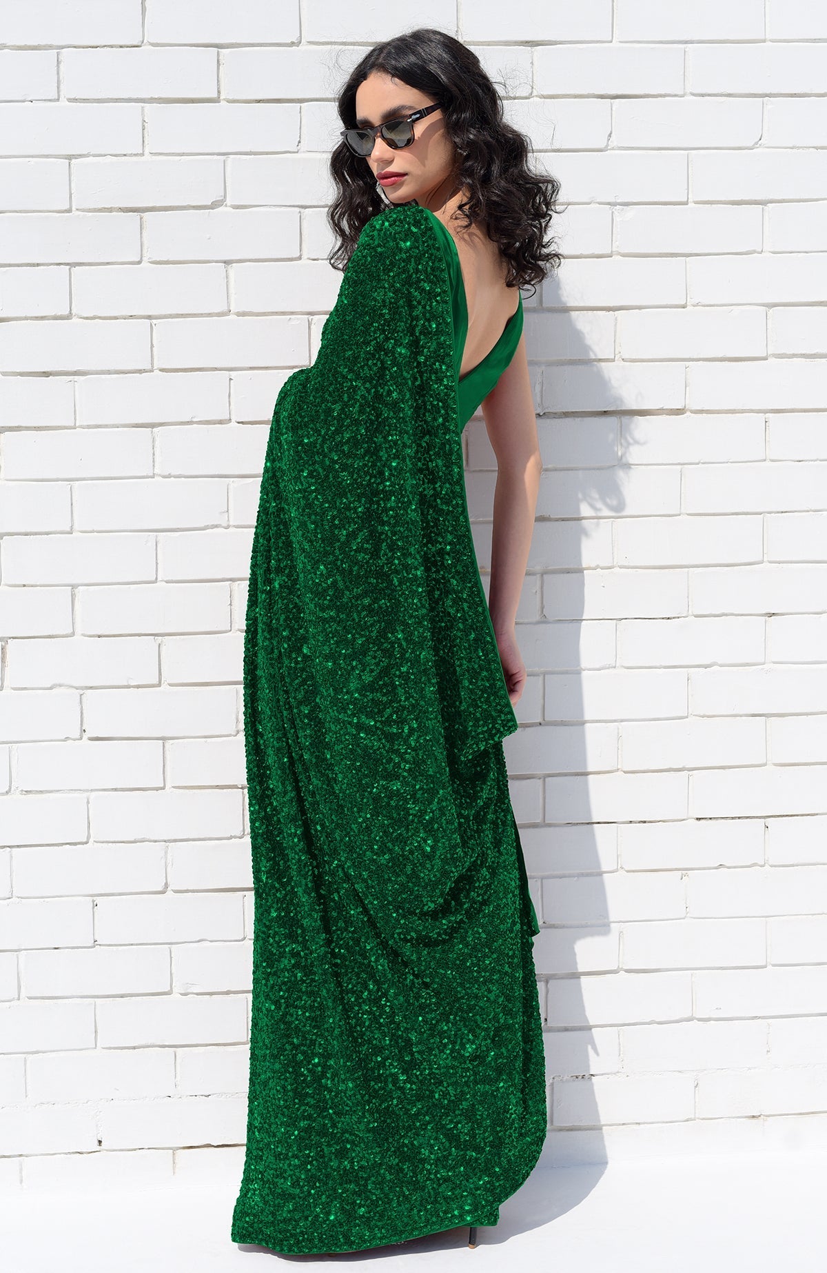 A woman in an emerald green sequin saree and sunglasses poses against a white brick wall.