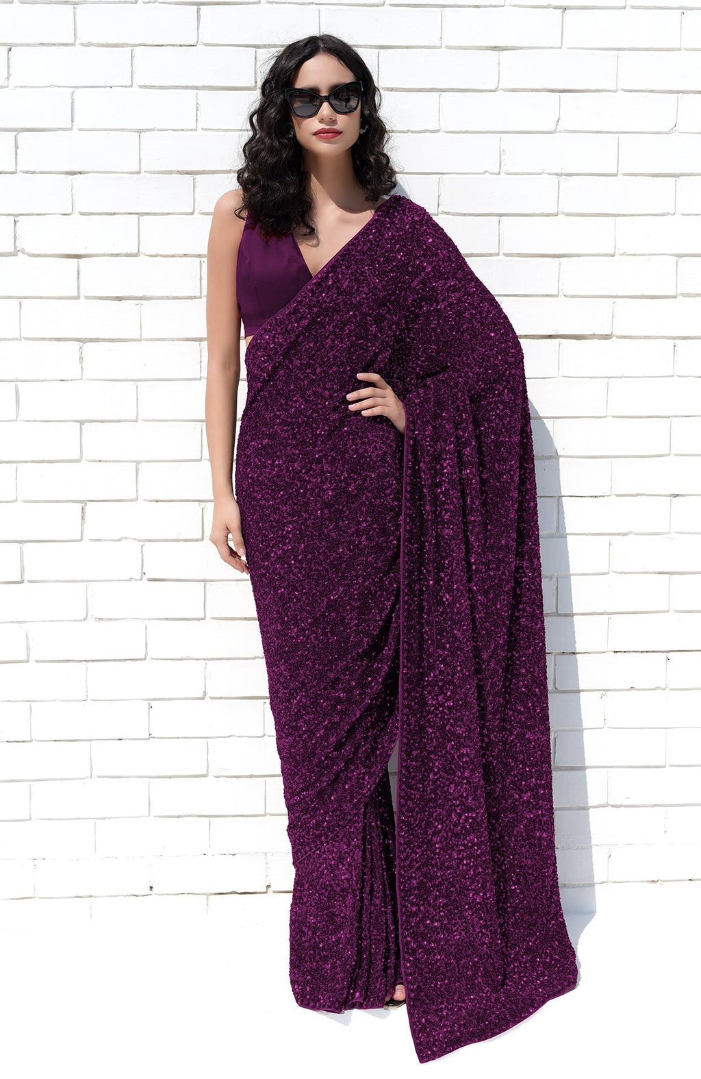 A woman in a purple sequin saree and matching blouse stands against a white brick wall.