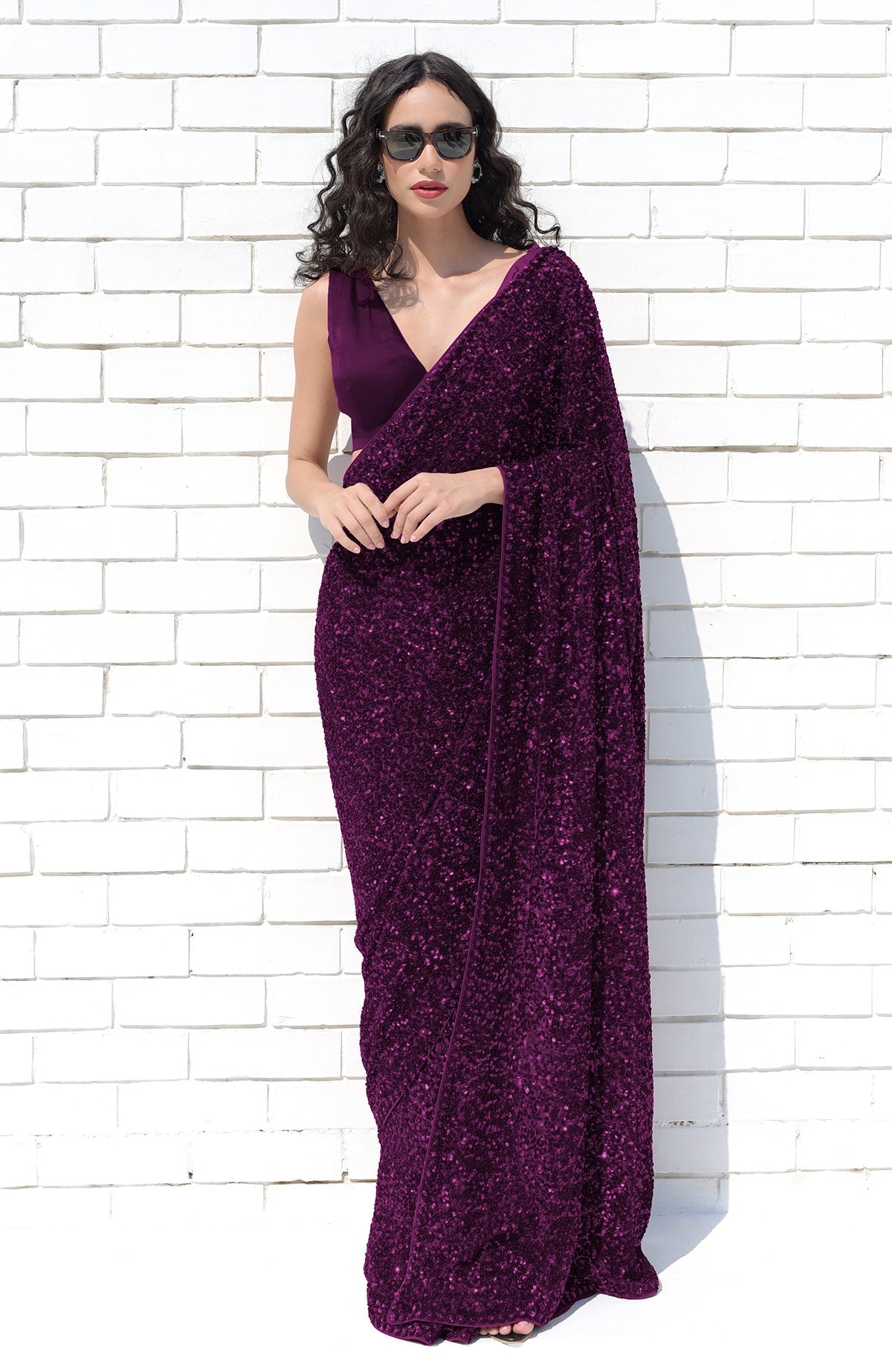 A woman in a deep purple sequin saree and matching blouse poses against a white brick wall.