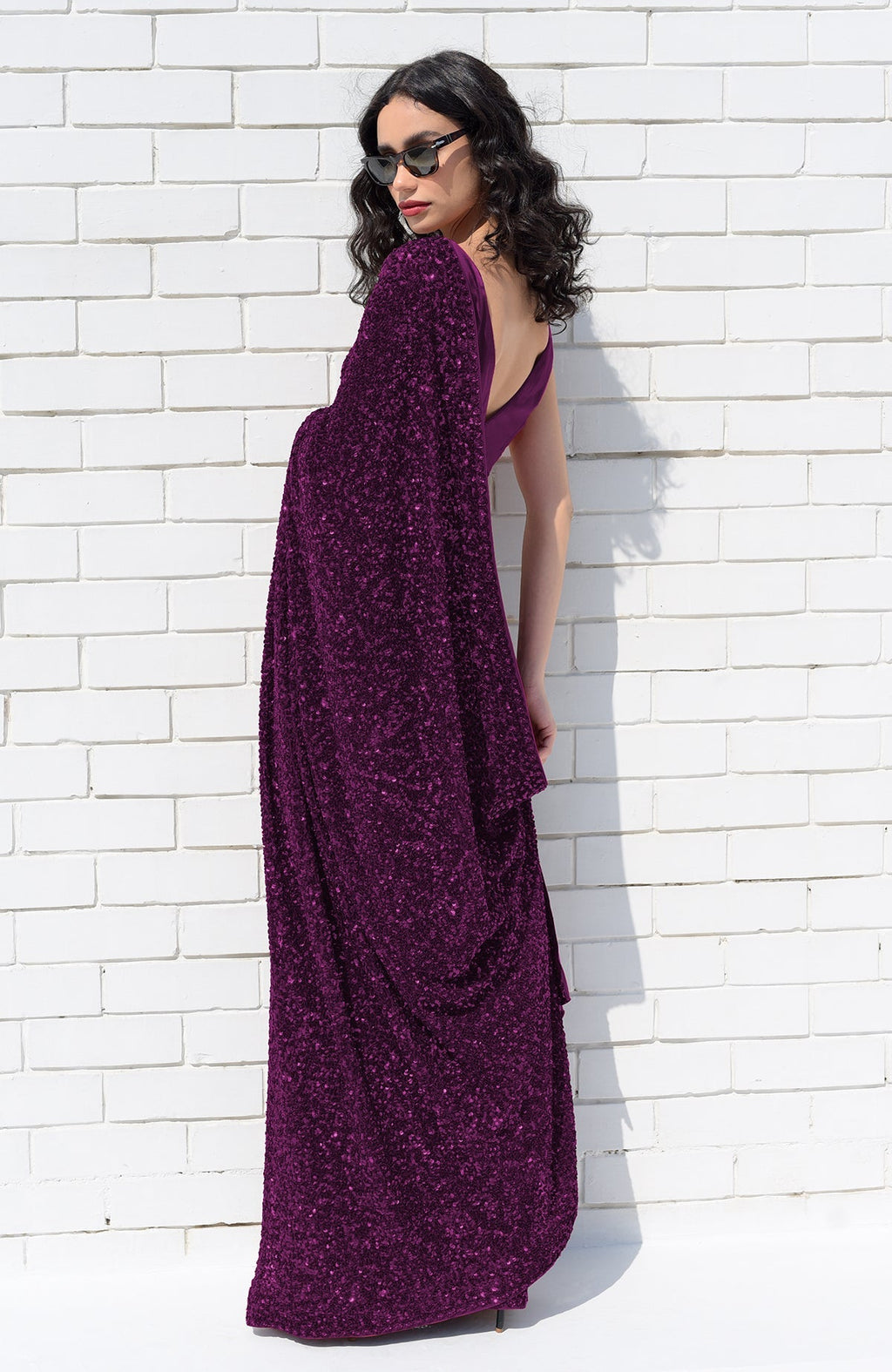 A woman in a purple sequin saree and sunglasses poses against a white brick wall.