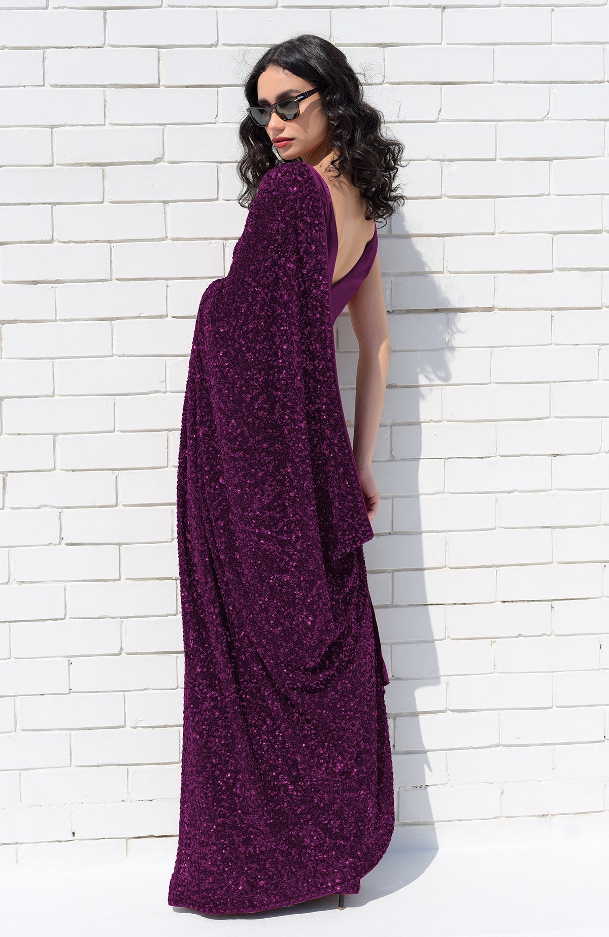 A woman in a purple sequin saree and sunglasses poses against a white brick wall.