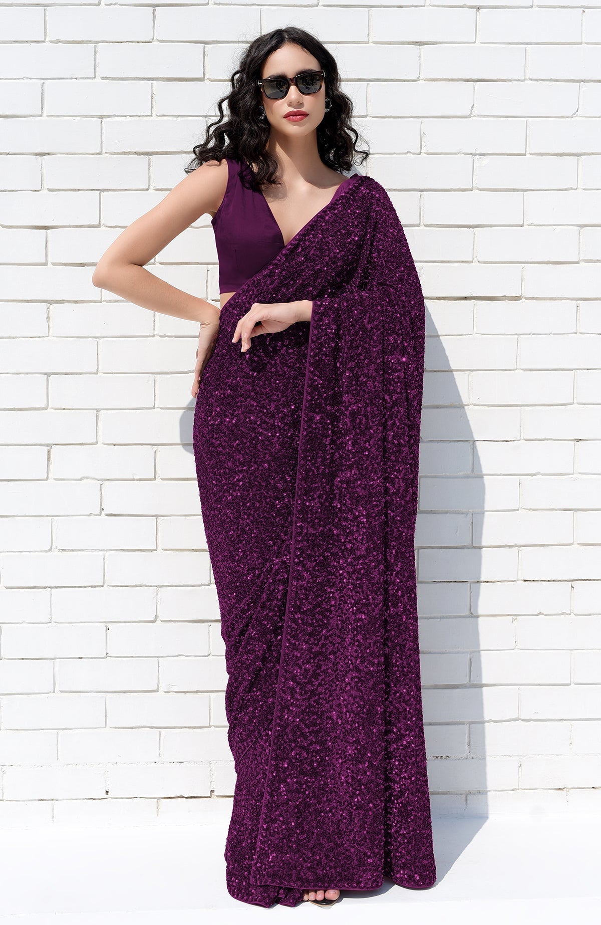 A woman in a purple sequin saree and matching blouse poses against a white brick wall.